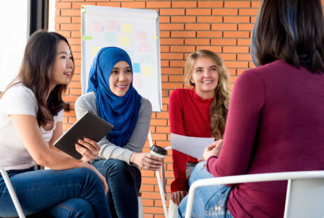 Group of multiethnic women, including a white woman, an Asian woman, and a woman in a hijab, have a discussion with a flipchart visible behind them