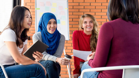 Group of multiethnic women, including a white woman, an Asian woman, and a woman in a hijab, have a discussion with a flipchart visible behind them