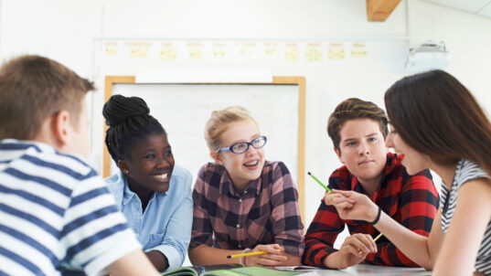 Group of diverse middle school students, including an African American girl, two white girls, and two white boys, having a discussion while writing